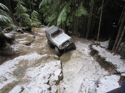 2008 Jeep Wrangler - An off-road vehicle descends a challenging trail with patches of snow and mud, surrounded by tall trees.