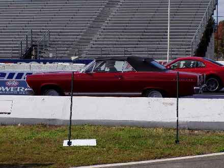 1999 Ford Crown Victoria - Two red cars, a convertible and a coupe, are lined up on a drag strip with grandstands.
