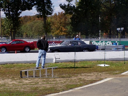 1999 Ford Crown Victoria - Two cars are lined up at a drag strip, with a person standing on a ladder between them.