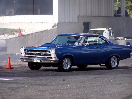 1999 Ford Crown Victoria - Blue 1966 Ford Fairlane coupe on a race track with a person and truck in the background.
