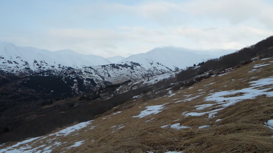 Snow-covered mountains rise above a dry, grassy slope with patches of snow under a cloudy sky