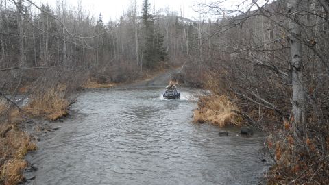 1986 Chevrolet Blazer - An ATV navigates a water crossing in a natural, wooded environment.