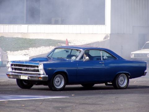 1999 Ford Crown Victoria - A blue 1967 Ford Fairlane coupe with a hood scoop is seen from the side, with another vehicle in the background.