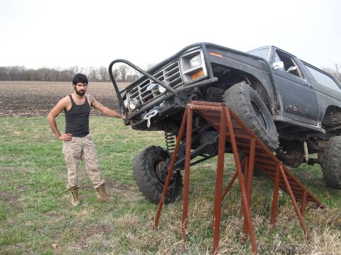 1986 Ford Bronco - A heavily modified SUV is positioned with its front tire on a ramp, while a man stands beside it.