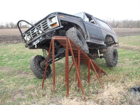 1986 Ford Bronco - Modified black Ford Bronco with oversized tires is positioned on a rusty metal ramp outdoors.