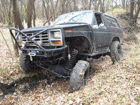 1986 Ford Bronco - Modified black SUV with a prominent brush guard and off-road tires navigates a wooded area.