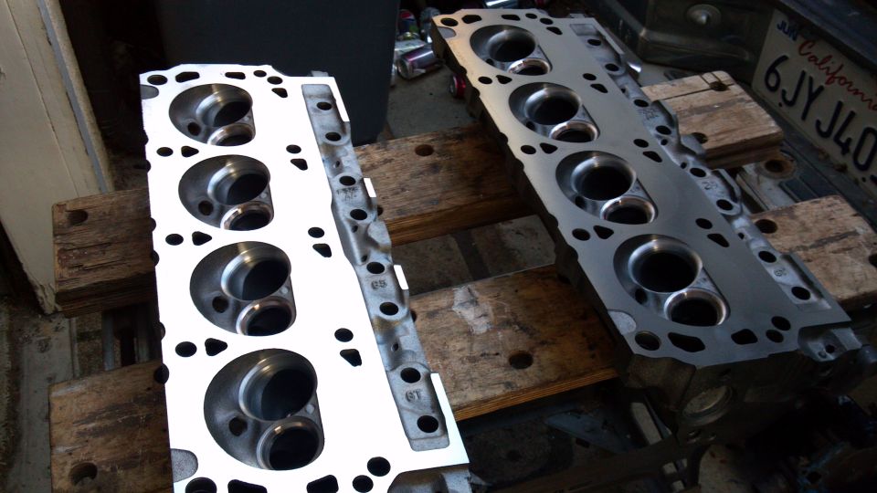 Two engine cylinder heads, one clean and white, the other metallic gray, resting on wooden pallets