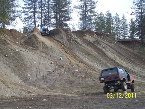 1988 Ford Bronco - Two SUVs navigate a steep dirt incline, one with a lifted suspension and exposed undercarriage.