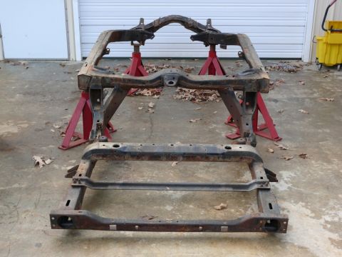 1976 Chevrolet Corvette - The chassis of a vehicle is supported by red jack stands on a concrete surface, with fallen leaves scattered around.
