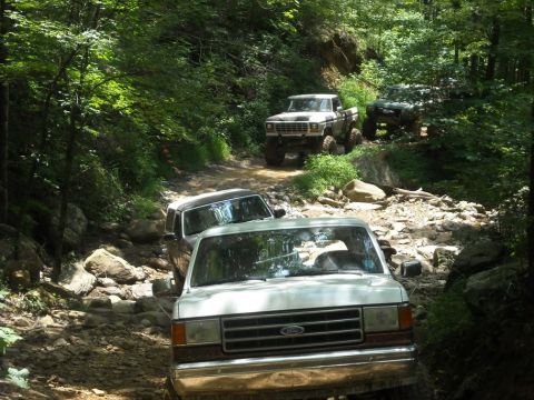 1989 Ford Bronco - Multiple trucks navigate a challenging rocky trail through a wooded area.