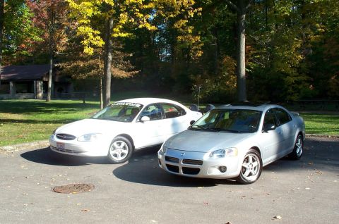 2001 Ford Taurus - A white 2001 Ford Taurus is parked next to a silver sedan in an outdoor setting with trees in the background.