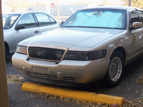 2000 Mercury Grand Marquis - A tan 2000 Mercury Grand Marquis is parked in front of another vehicle, showing its front grille and custom wheels.