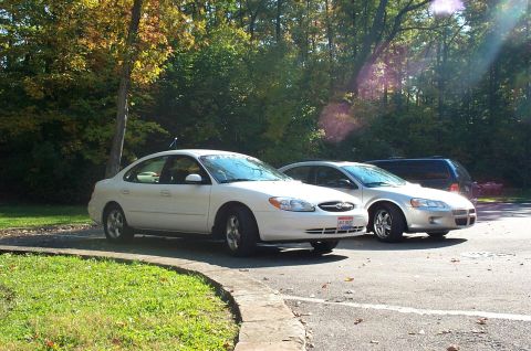 2001 Ford Taurus - Two sedans are parked in a lot with trees in the background.
