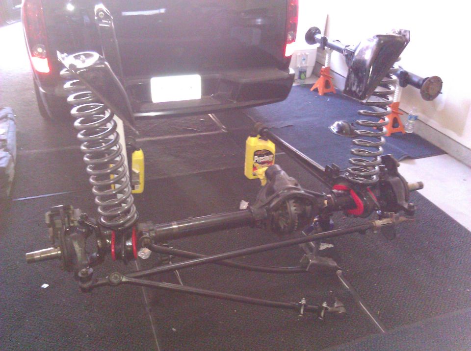 Disassembled front axle, coil springs, and suspension parts laid out on the floor, with the rear of a black SUV visible in the background