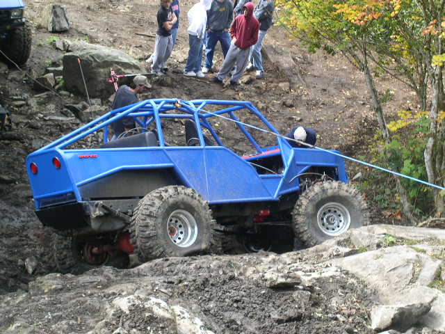 Blue caged Bronco trail rig being winched up a muddy rock face with spectators
