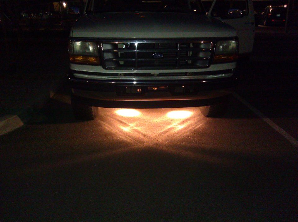 White Ford truck front view, grille and headlights visible, with underglow lights illuminating the ground
