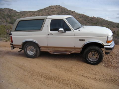 1995 Ford Bronco - A white 1995 Ford Bronco with tan accents is shown from the front three-quarter view on a dusty road with hills in the background.