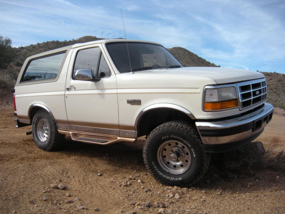White Ford Bronco parked on dirt road with hills in background. Has chrome accents and running boards