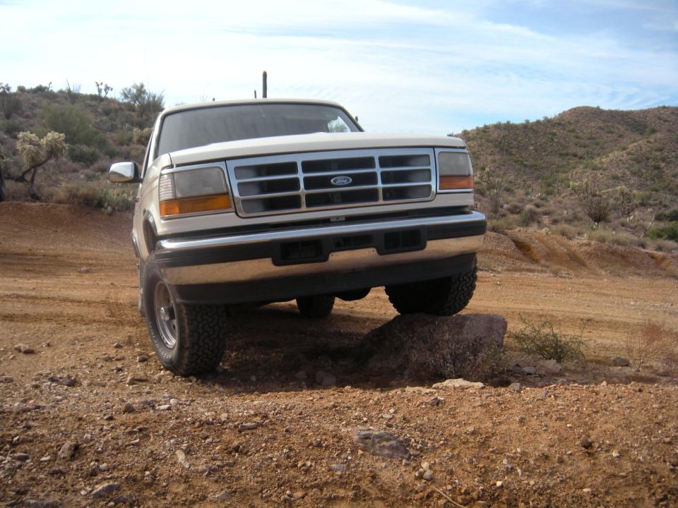 White Ford Bronco with chrome grille, front bumper, and side mirror, driving on a rocky, desert terrain