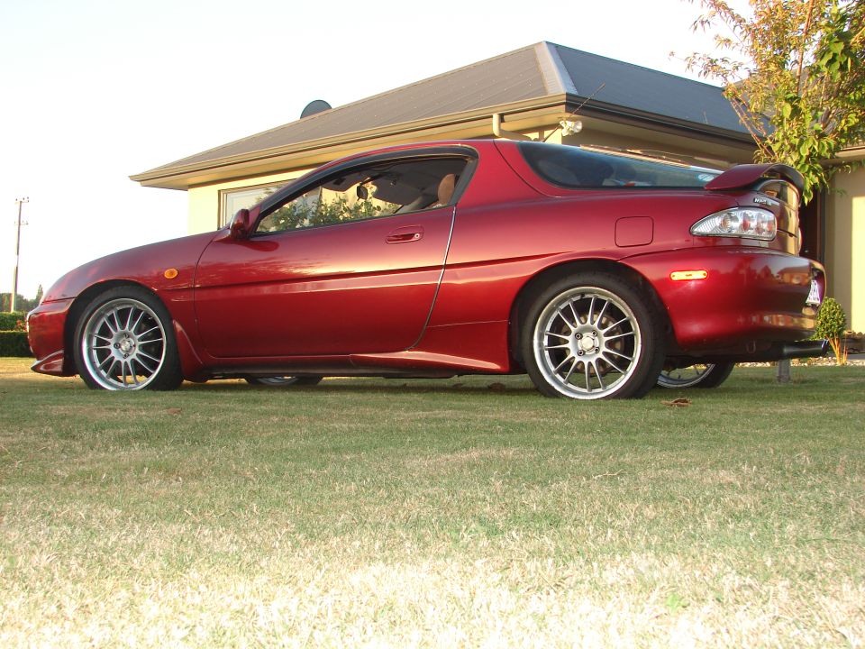 Side view of a red coupe with aftermarket wheels parked on a lawn in front of a house
