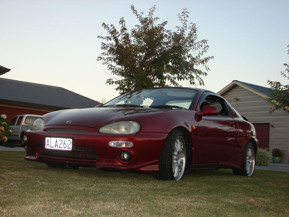 Red 1993 Mazda MX-3 parked on lawn in front of a house and garage. License plate ALA262