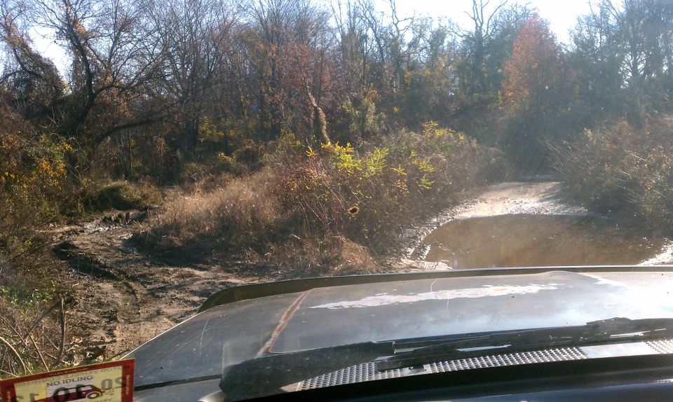 View from inside a vehicle driving on a muddy trail with trees and brush on either side