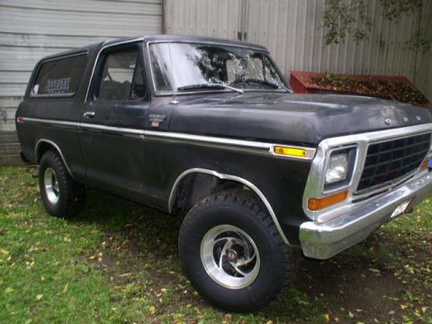 1978 Ford Bronco - Faded flat-black late-1970s Ford Bronco with hardtop parked on grass beside a corrugated metal building, wearing polished five-spoke wheels…