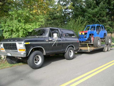 1978 Ford Bronco - A black first-generation-style full-size Ford Bronco SUV parked at an angle on a two-lane road, towing a flatbed trailer that carries a blu…