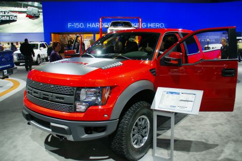 1993 Ford Taurus - A red Ford F-150 Raptor is displayed with its door open at an auto show, with other vehicles and signage in the background.