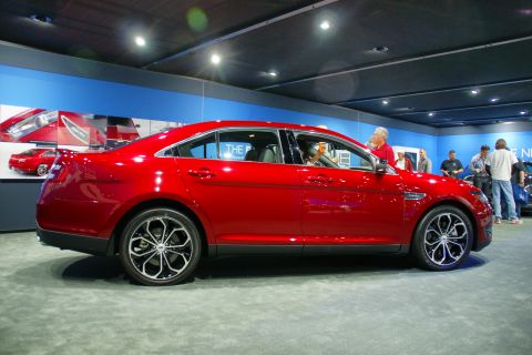 1993 Ford Taurus - A red sedan is displayed indoors with people observing it.