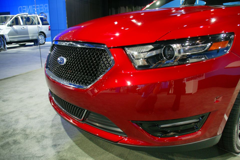 Close-up of a red Ford sedan's front grille and headlight. Another silver SUV is visible in the background