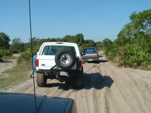 1995 Ford Bronco - Two SUVs drive down a sandy dirt road surrounded by trees and brush.