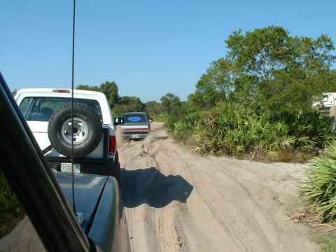 1995 Ford Bronco - Two SUVs travel down a dusty, unpaved road surrounded by dense vegetation under a clear blue sky.