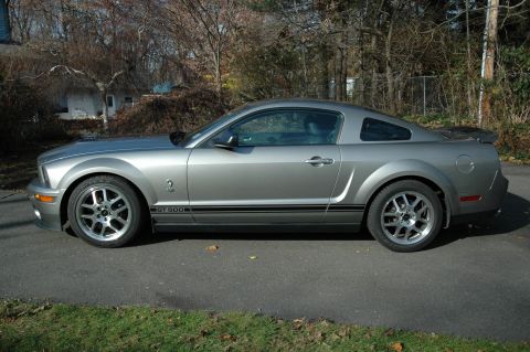 2008 Ford Mustang - Side profile of a silver 2008 Ford Mustang GT 500 parked on an asphalt driveway with grass and trees in the background.