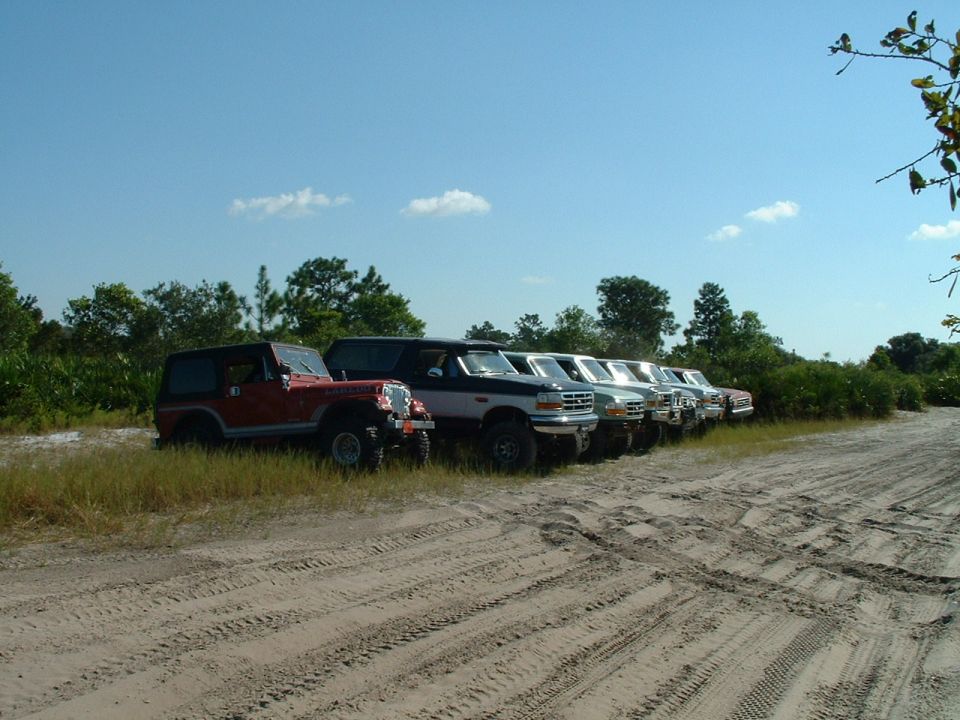 Several SUVs and trucks parked in a line on a dirt road with trees and blue sky in the background