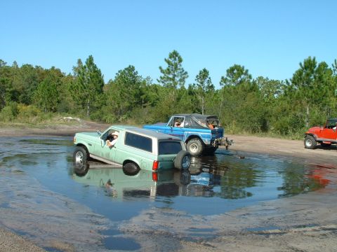 1995 Ford Bronco - A light green 1995 Ford Bronco is stuck in a muddy puddle, with a blue Jeep and a red Jeep nearby.