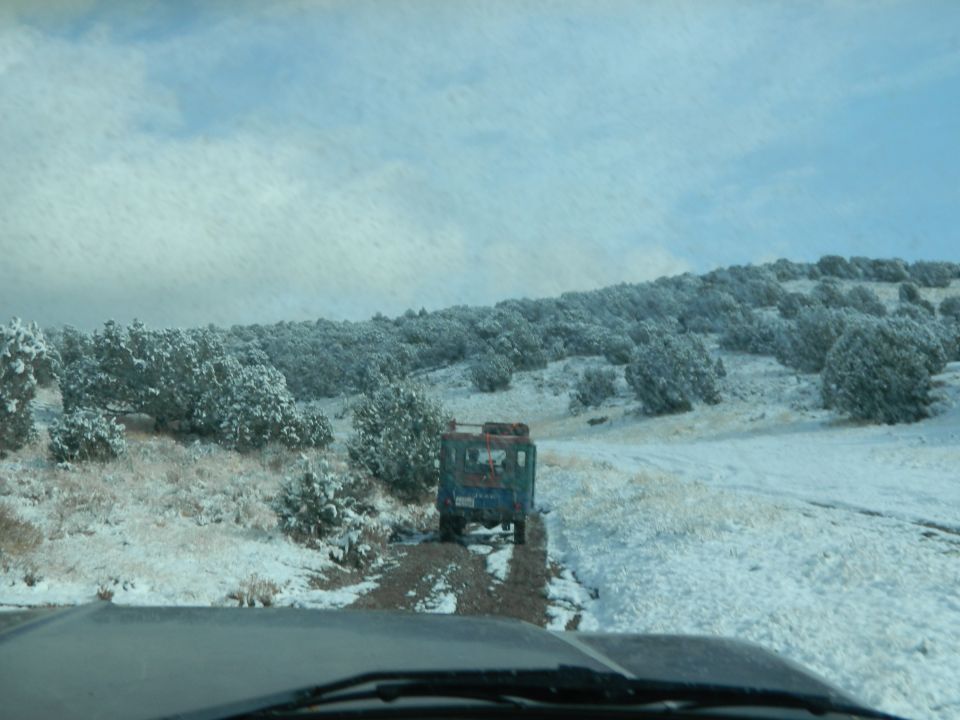 Blue off-road vehicle driving on a dirt road in a snowy landscape. Another vehicle is visible in the foreground