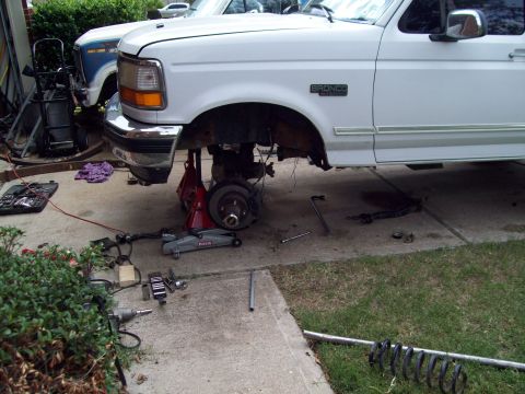1996 Ford Bronco - Front wheel removed from a white 1996 Ford Bronco undergoing lift installation. Tools and jacks are visible around the vehicle.