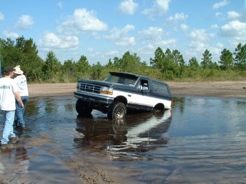 1995 Ford Bronco - A two-tone Ford Bronco is partially submerged in water on a sunny day with clouds in the sky.