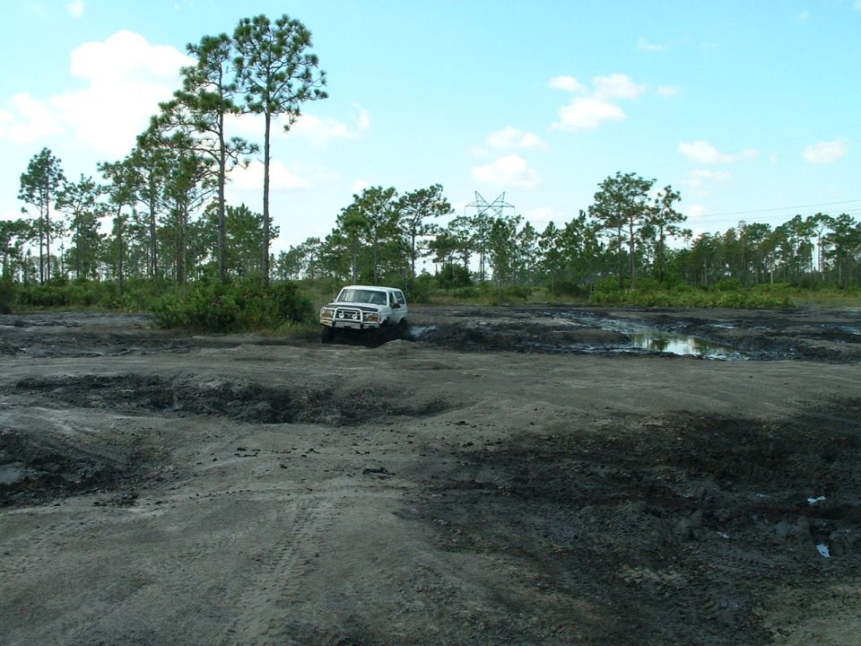 1995 Ford Bronco driving through a muddy field with trees in the background