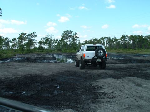 1995 Ford Bronco - A white Ford Bronco navigates a muddy, off-road area under a partly cloudy sky.