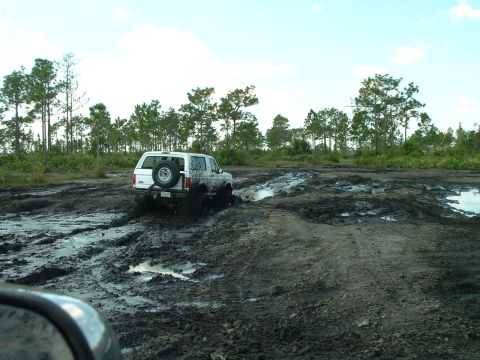 1995 Ford Bronco - White 1995 Ford Bronco navigates a muddy off-road course, kicking up dirt and water.