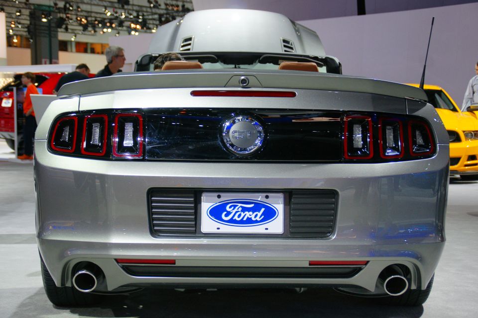 Rear view of a silver Ford Mustang convertible with its top up, showing taillights and a Ford license plate frame. A yellow car is visible to the right
