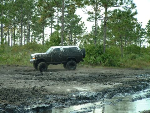 1995 Ford Bronco - A black 1995 Ford Bronco navigates a challenging muddy terrain, kicking up dirt as it goes.
