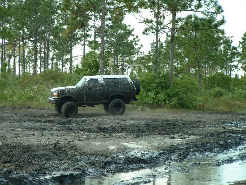 Muddy 1995 Ford Bronco driving through a muddy off-road course with trees in the background