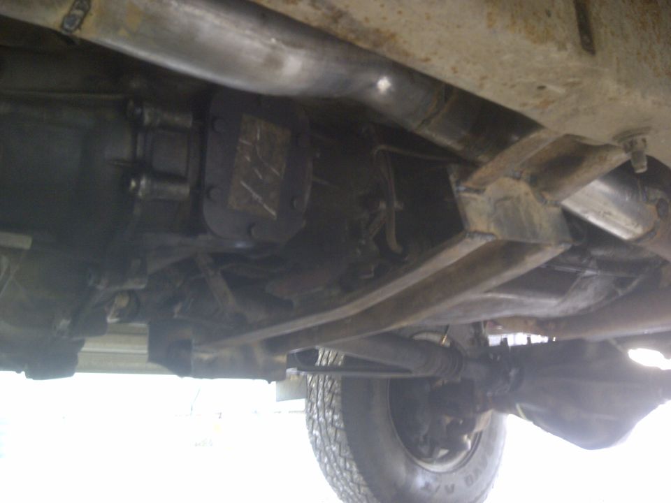 Underside of a vehicle with visible transmission, driveshaft, and rear axle. A tire is partially visible on the right