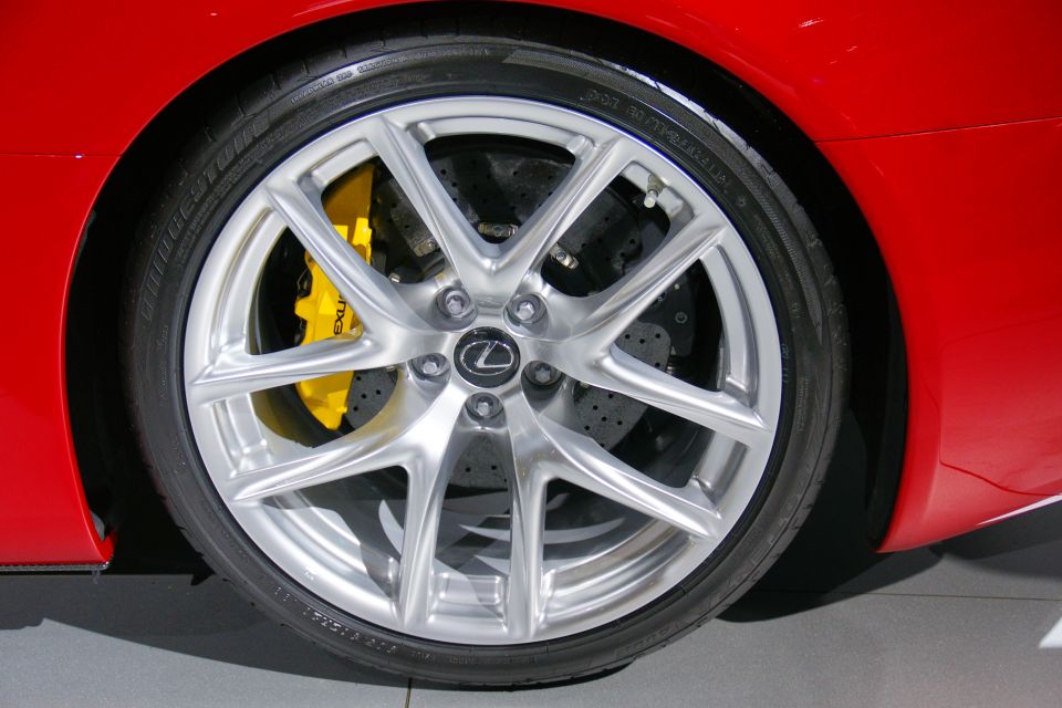 Close-up of a red car's front wheel with a silver multi-spoke rim and yellow brake caliper. The Lexus logo is visible on the wheel center cap