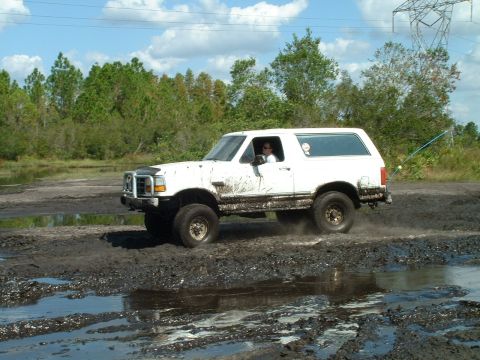 1995 Ford Bronco - White 1995 Ford Bronco navigates a muddy track with trees and power lines in the background.