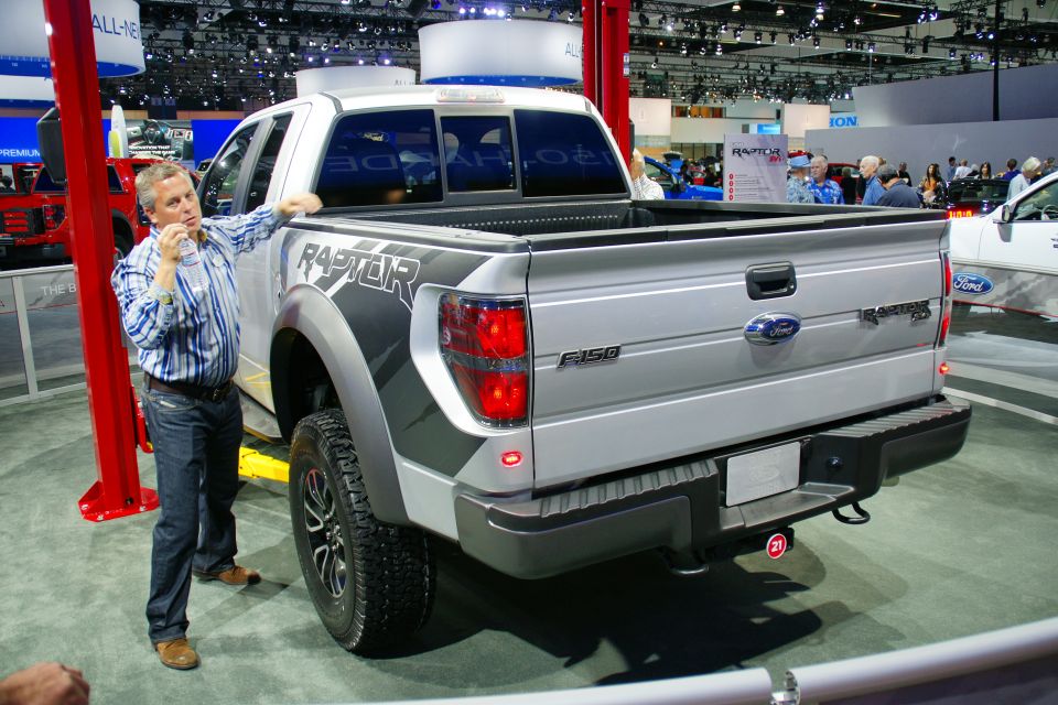 Rear view of a silver Ford F-150 Raptor pickup truck with 'RAPTOR' graphics on the fender. A man stands next to it