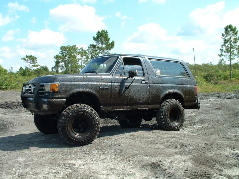 1995 Ford Bronco - Mud-covered 1995 Ford Bronco sits on a dirt track under a partly cloudy sky.